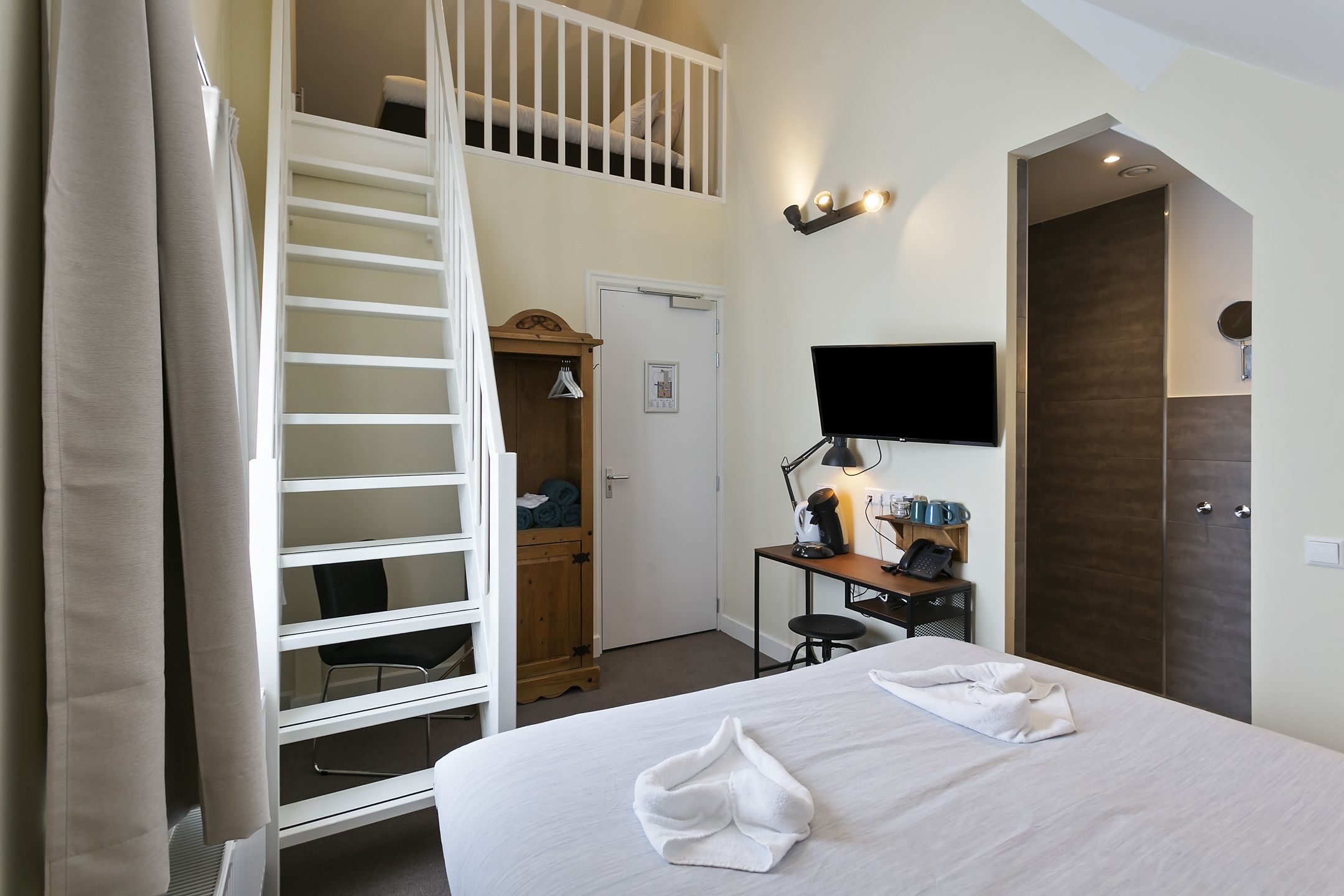 A hotel room with a bed, a desk with a computer, a chair, a wardrobe, and a staircase to a loft.
