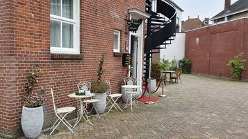Een bakstenen gebouw met een wenteltrap, terras met witte metalen stoelen en tafels, en potplanten.