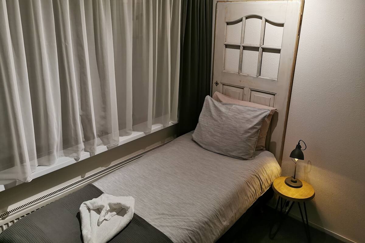 A bedroom with a bed, a bedside table with a lamp, a window with sheer curtains, and a wall-mounted artwork.