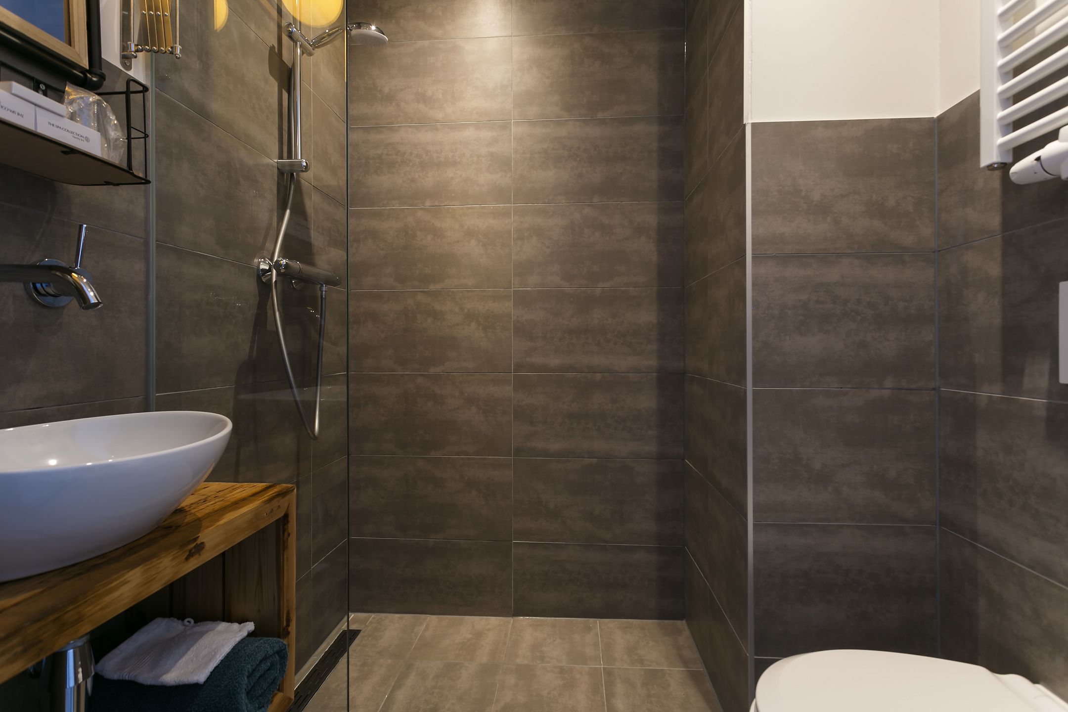 A modern bathroom with a large white sink, a wooden shelf, a glass shower enclosure, and a toilet.