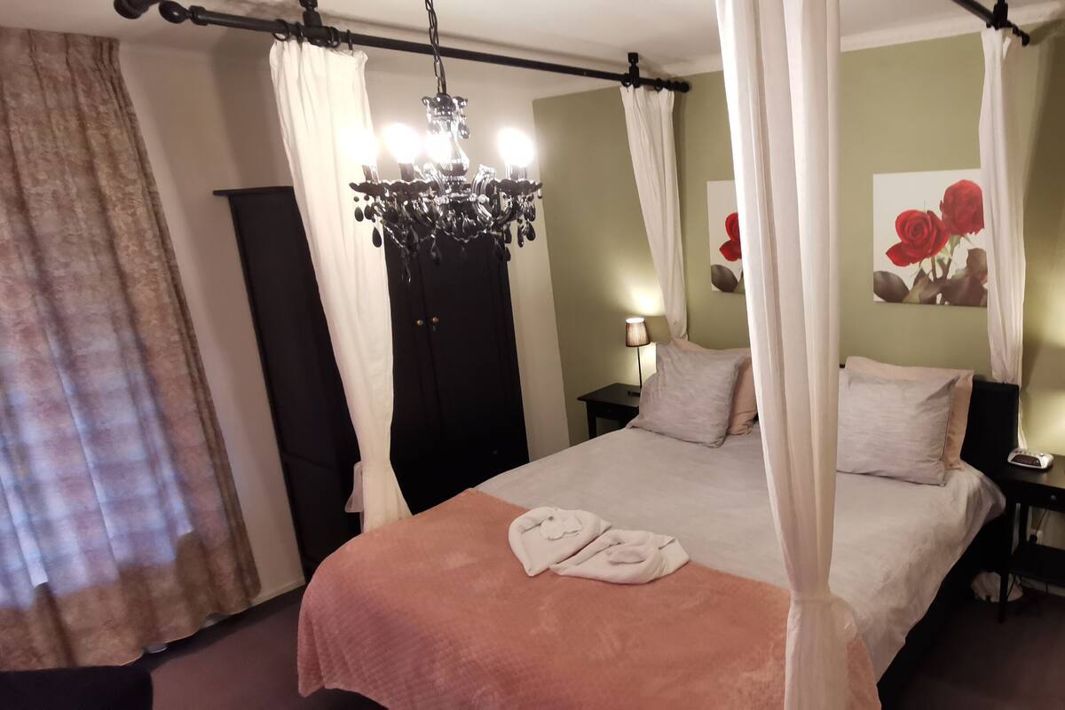 A bedroom with a canopy bed, two bedside lamps, a bedside table with a lamp, a painting of roses, and a chandelier.