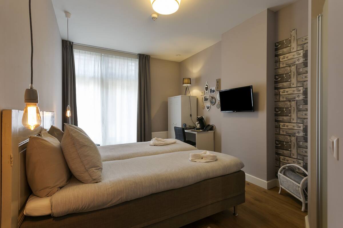 A hotel room with two beds, a desk with a computer, a TV, and a wall with suitcases.