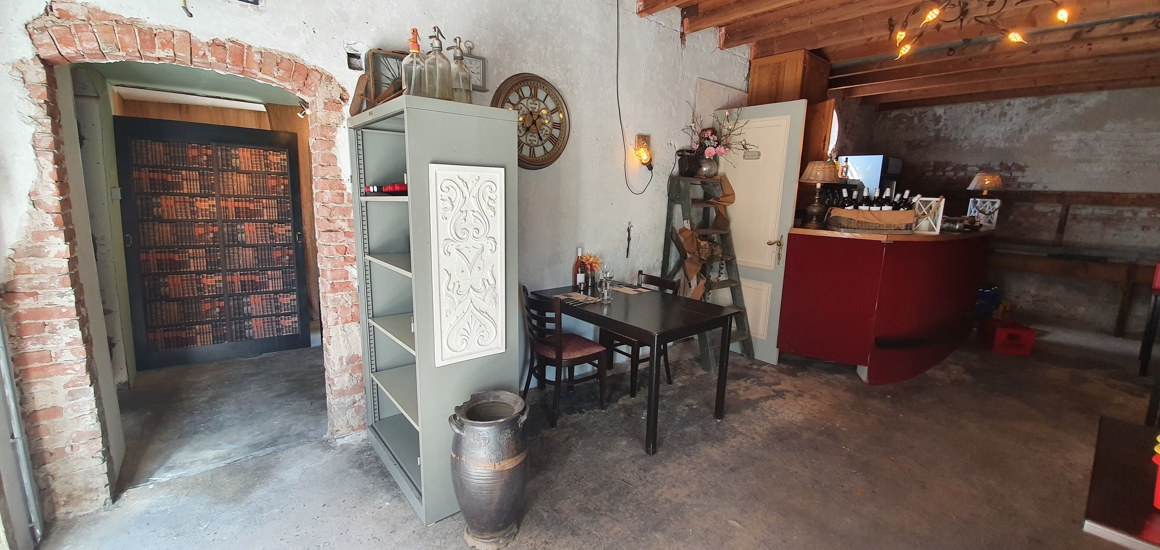 Een eenvoudige kamer met een bakstenen muur, een houten tafel met stoelen, een boekenkast, een rode kast en een decoratieve klok.