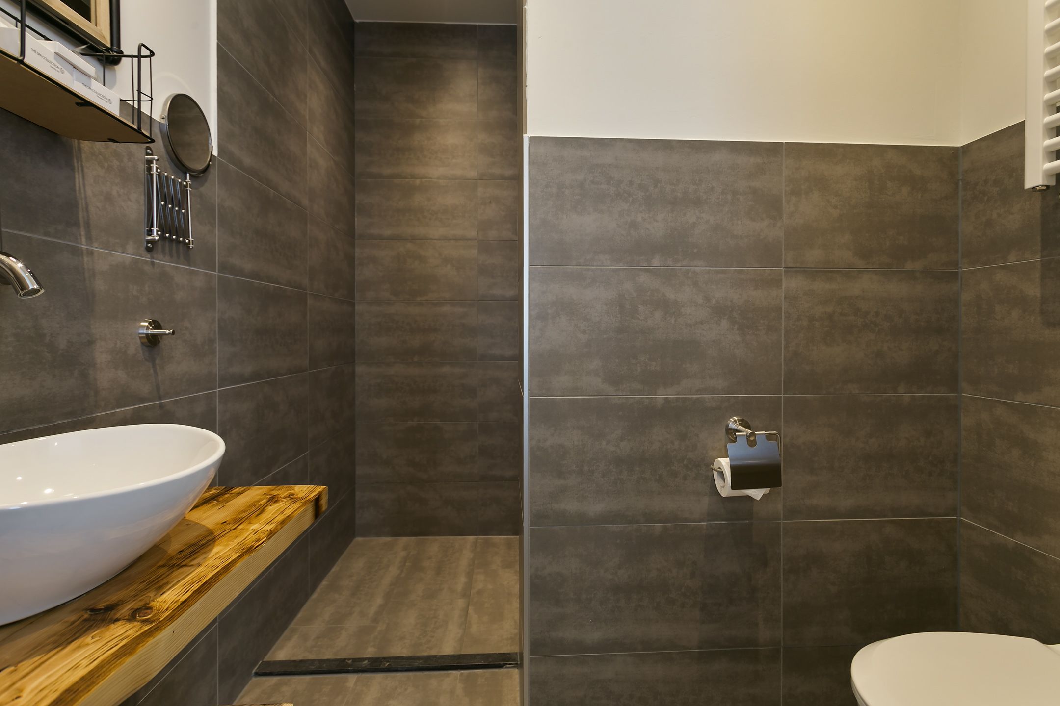 A modern bathroom with a large white sink, a wooden vanity, a toilet, and a shower area with dark tiles.