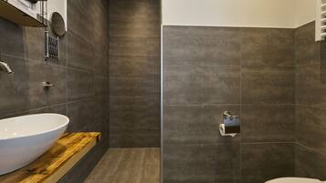 A modern bathroom with a large white sink, a wooden vanity, a toilet, and a shower area with dark tiles.