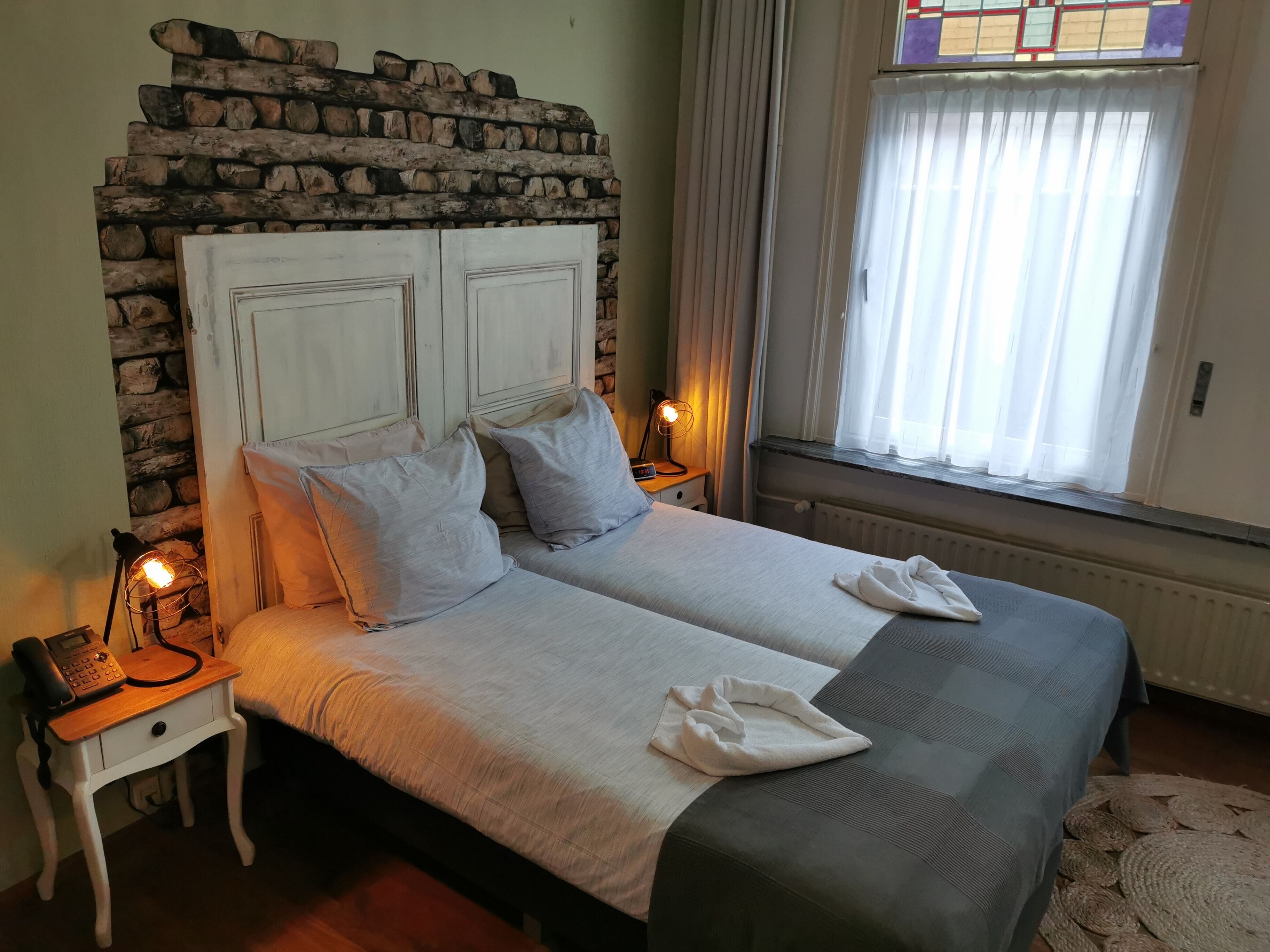 Uma cama de casal com cabeceira de parede de pedra, luminárias laterais e janela com cortinas.