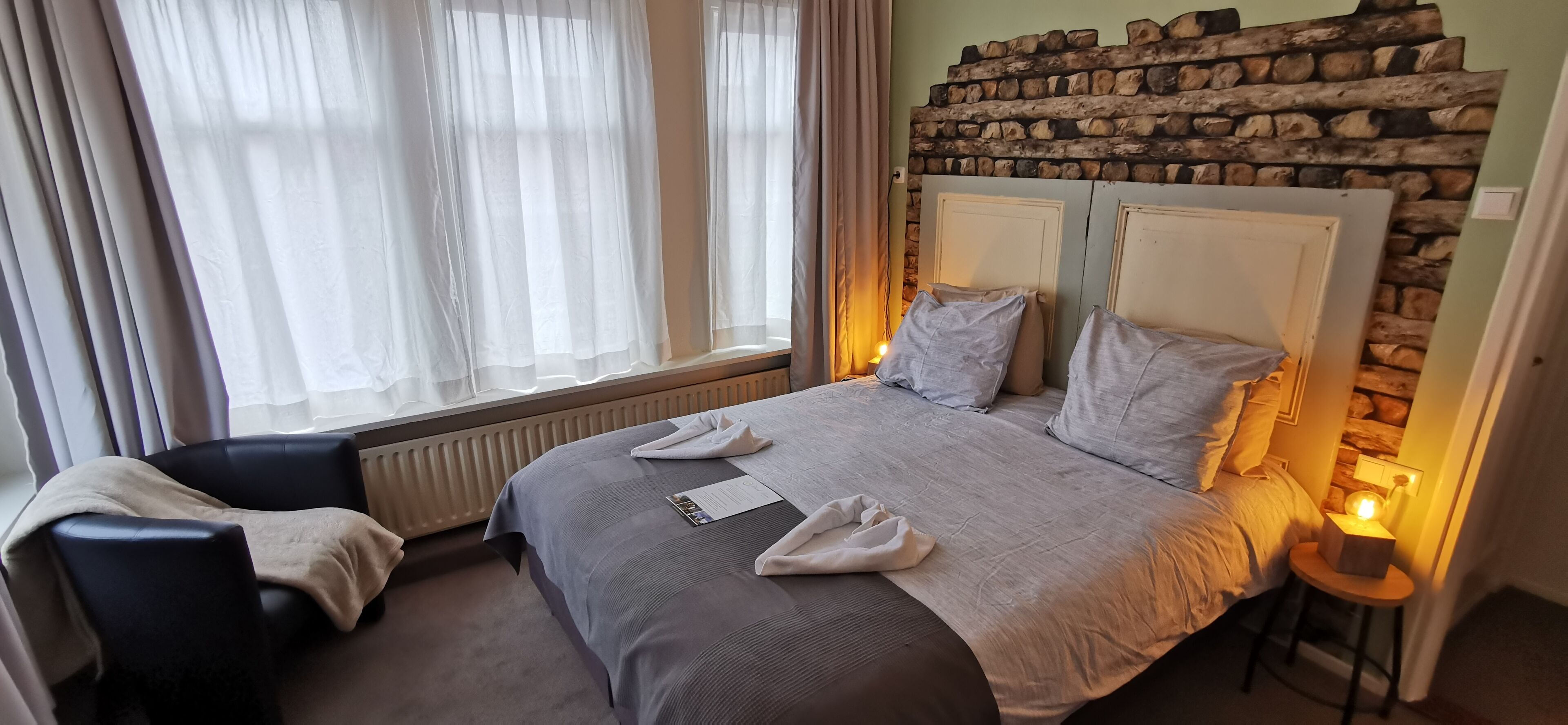 Una habitación de hotel con una cama grande, una silla, cabecera de madera y una ventana con cortinas translúcidas.