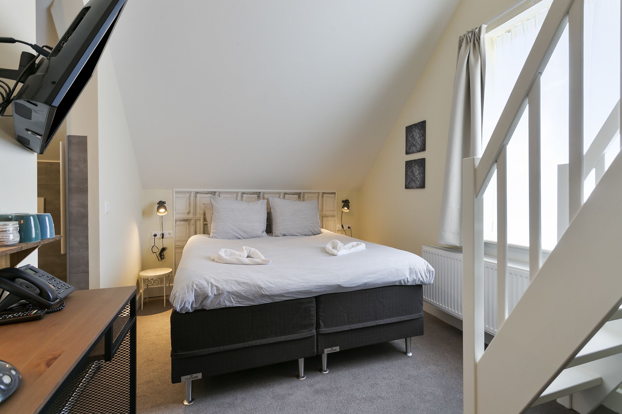 A bedroom with a bed, bedside lamps, a desk with a telephone, and a window with curtains.