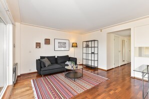 A modern living room with a dark grey sofa, a round coffee table, and a striped rug. There is a bookshelf with books and decorative items, and a framed picture on the wall.