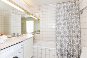 A bathroom with a white tiled wall, a bathtub, a shower curtain with a floral pattern, a sink with a mirror, and a washing machine.