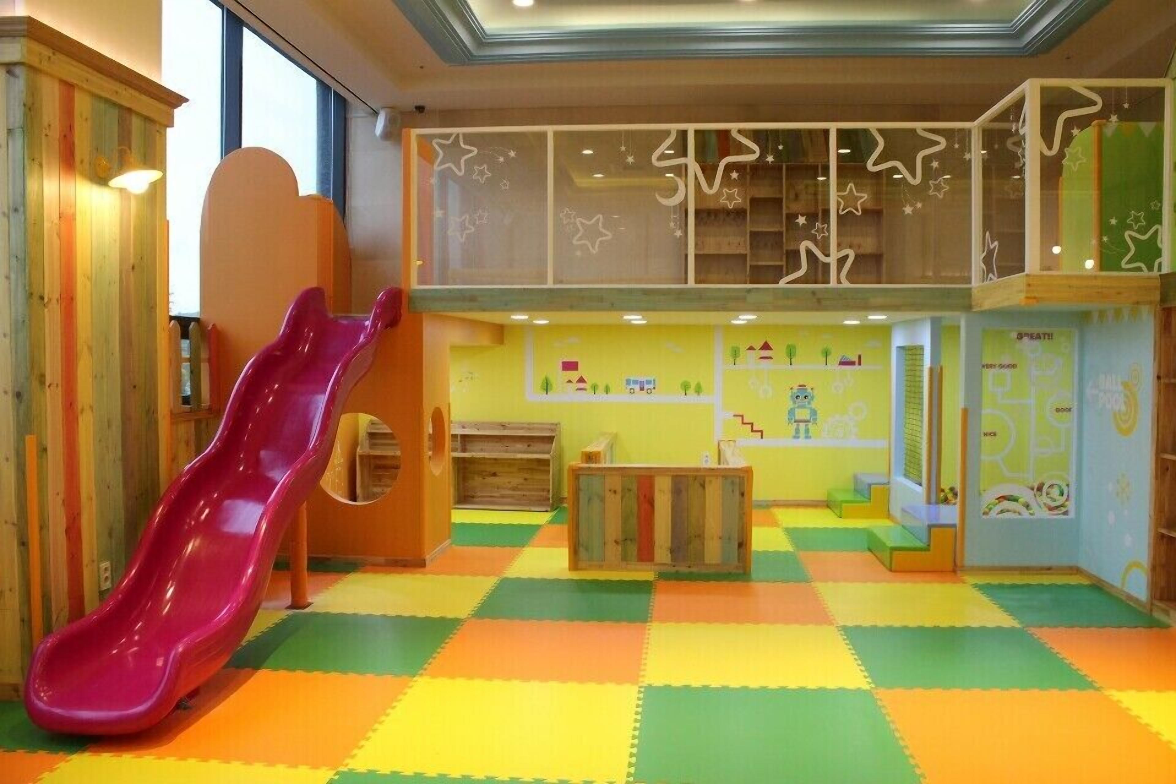 Un espace de jeux pour enfants avec un toboggan rose, des tapis de jeu colorés et des éléments décoratifs muraux à vocation éducative.