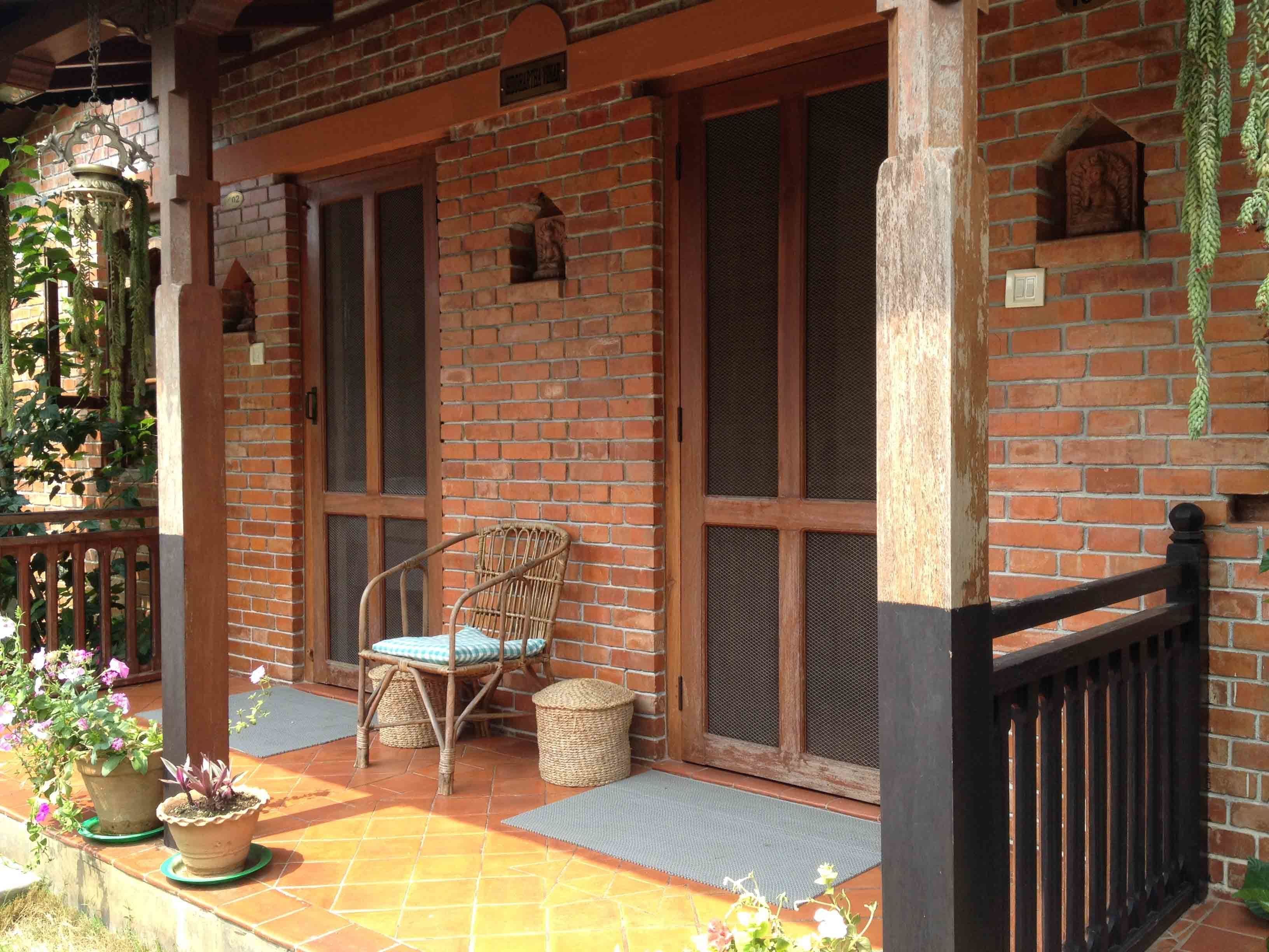A covered porch with a wicker chair and potted plants, brick walls, and a black railing.