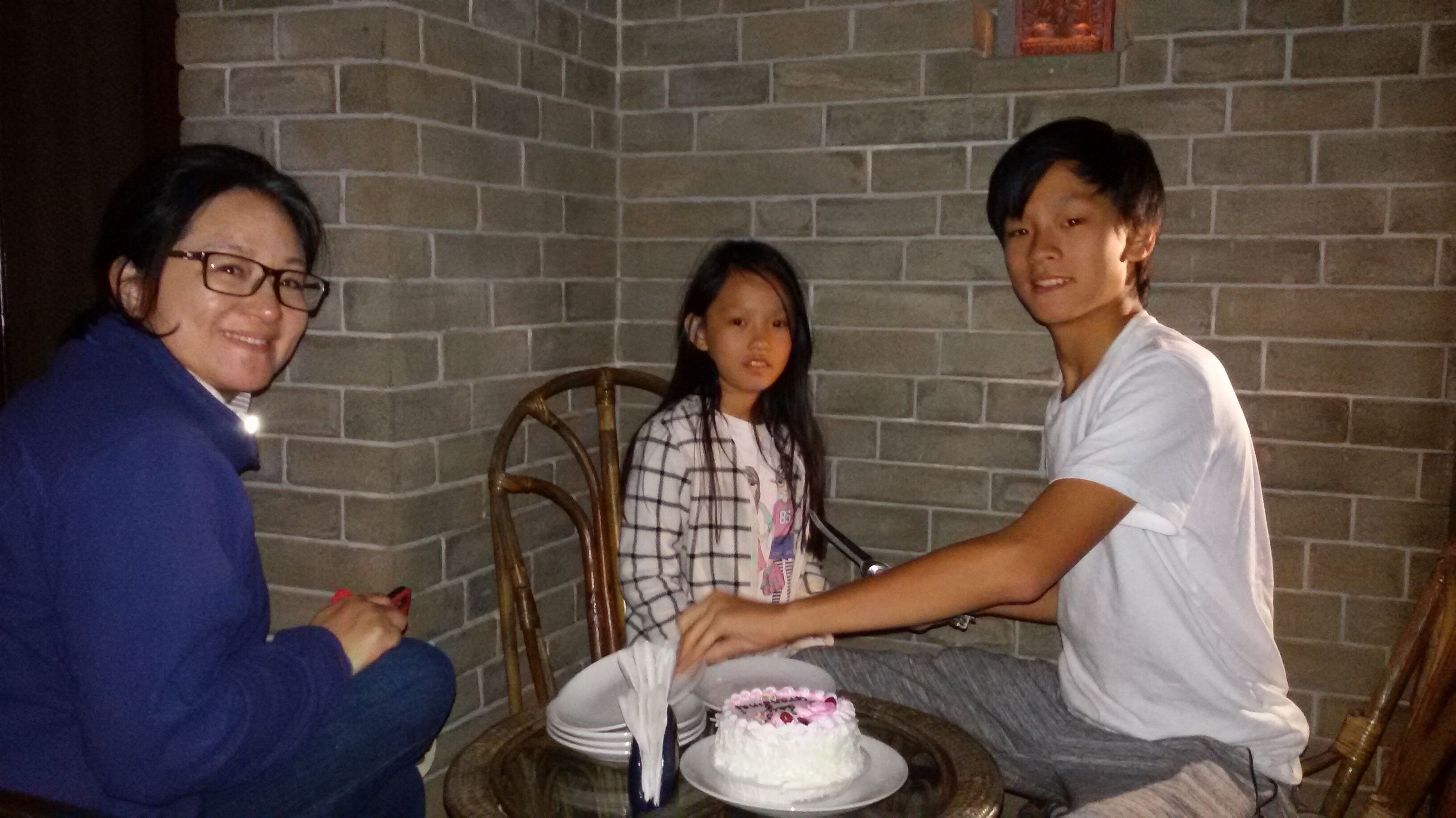 Three people seated at a table with a cake, in a room with brick walls.