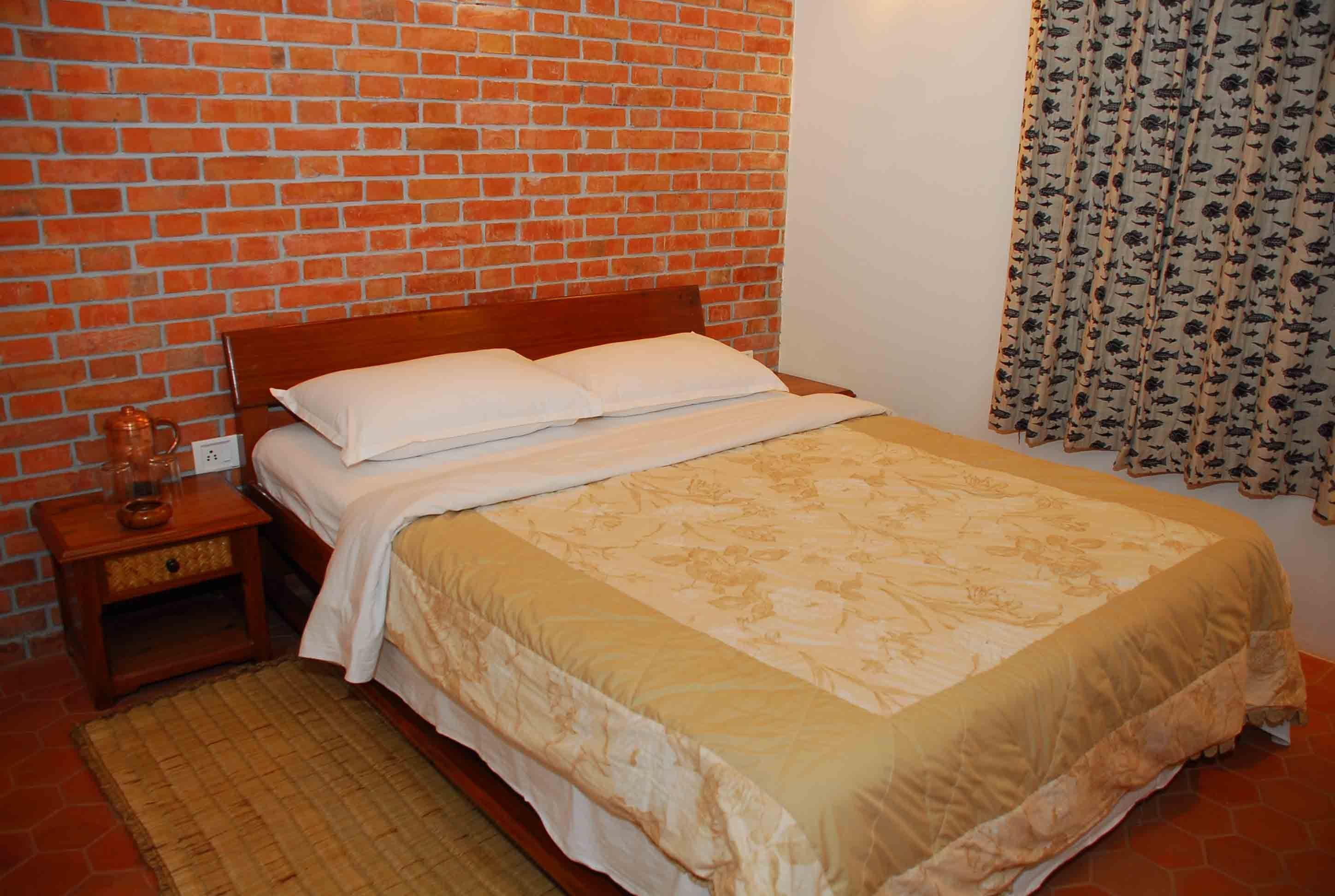 A bedroom with a brick wall, a wooden bed frame, a beige bedspread, a nightstand with a lamp, and a patterned curtain.
