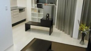 Una sala de estar moderna con un sofá oscuro, una mesa de centro y un televisor. Hay una zona de cocina con armarios blancos y un microondas.