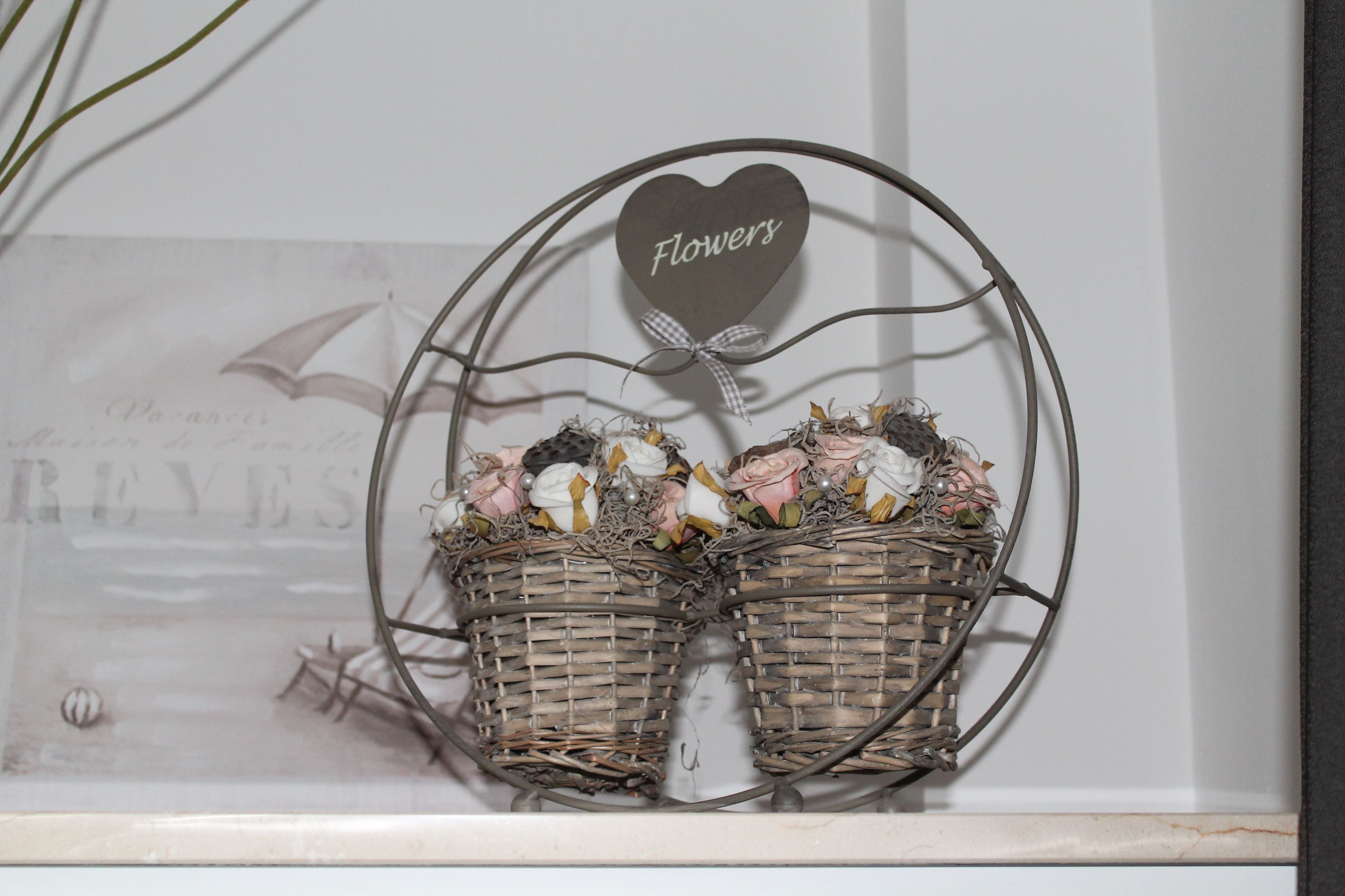Un panier en métal décoratif, orné d’une étiquette en forme de cœur sur laquelle est inscrit le mot « Fleurs », ainsi que deux paniers en osier remplis de fleurs séchées.