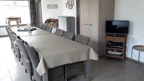 A spacious dining area with a long table and wicker chairs, a television on a wooden stand, and a wardrobe in the background.