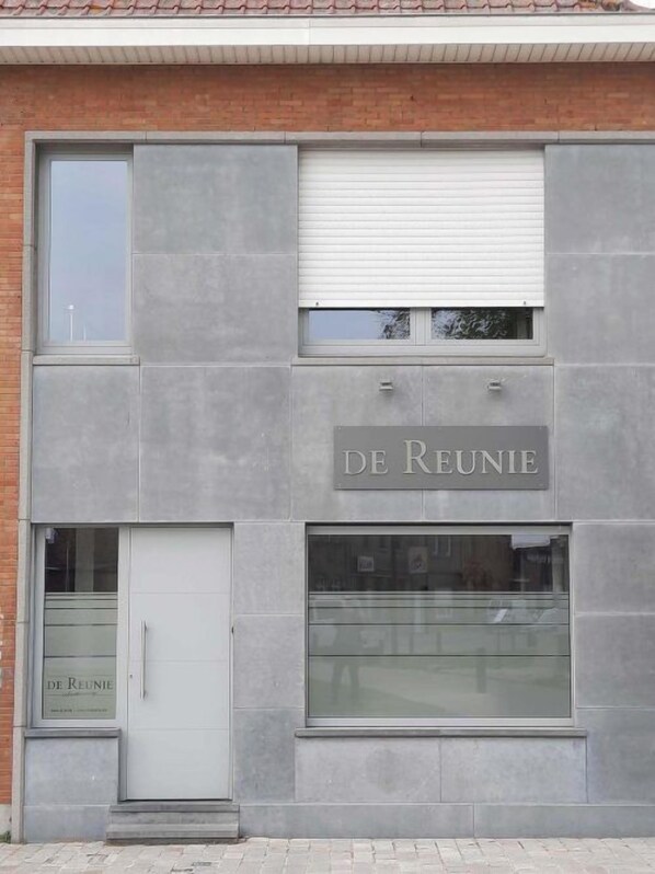 A building with a grey facade, a sign reading "DE REUNIE", and a glass door entrance.