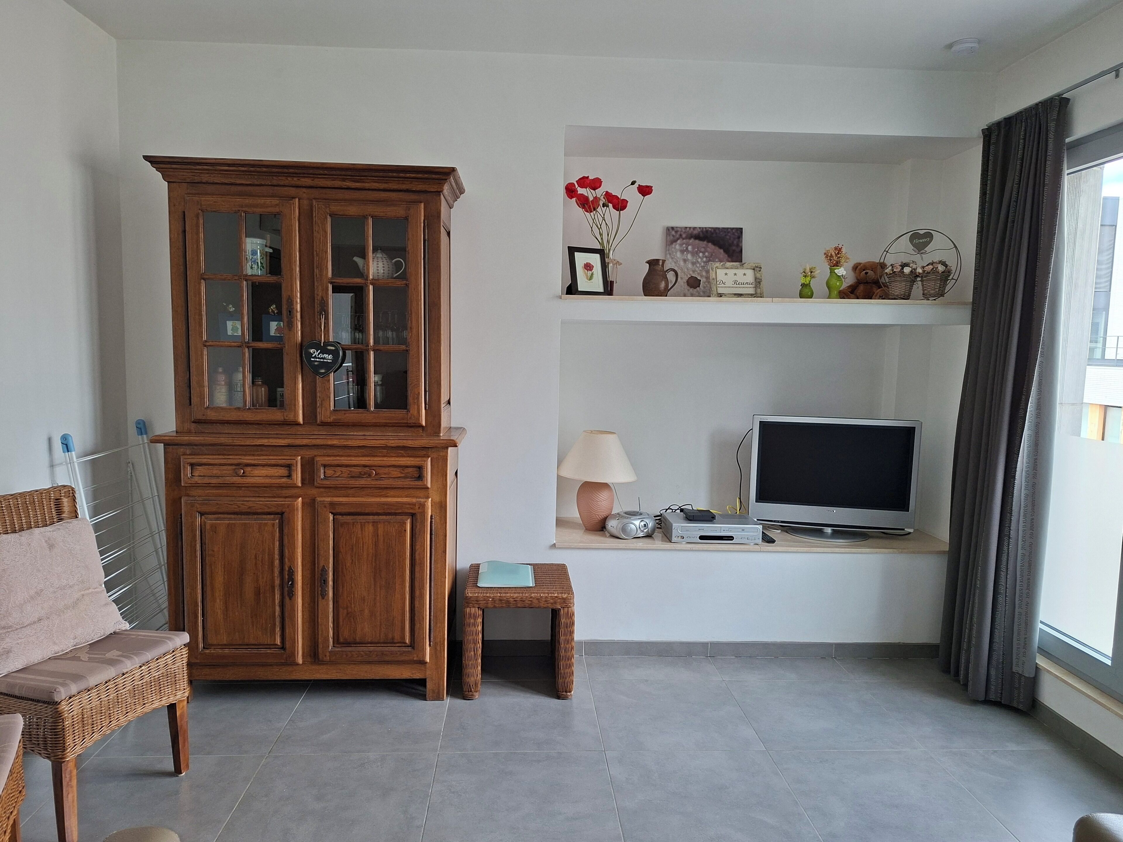 A living room with a wooden cabinet, a television, a small table, a wicker chair, and a window with curtains.
