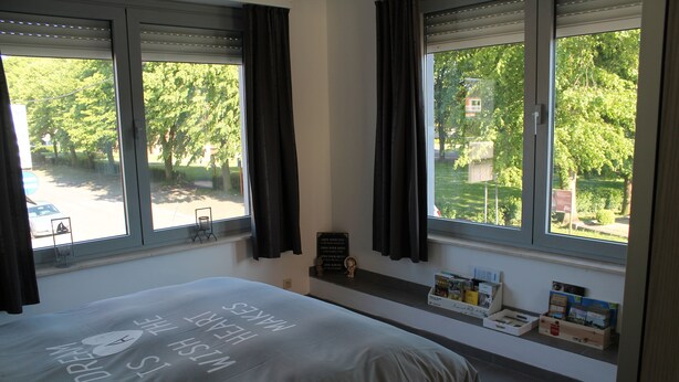 A bedroom with a bed, large windows, a shelf with books and toiletries, and a view of trees and a street.