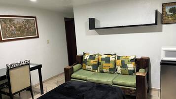 Una habitación con un sofá verde, un otoman negro, una silla de madera, una mesita y un cuadro en la pared.