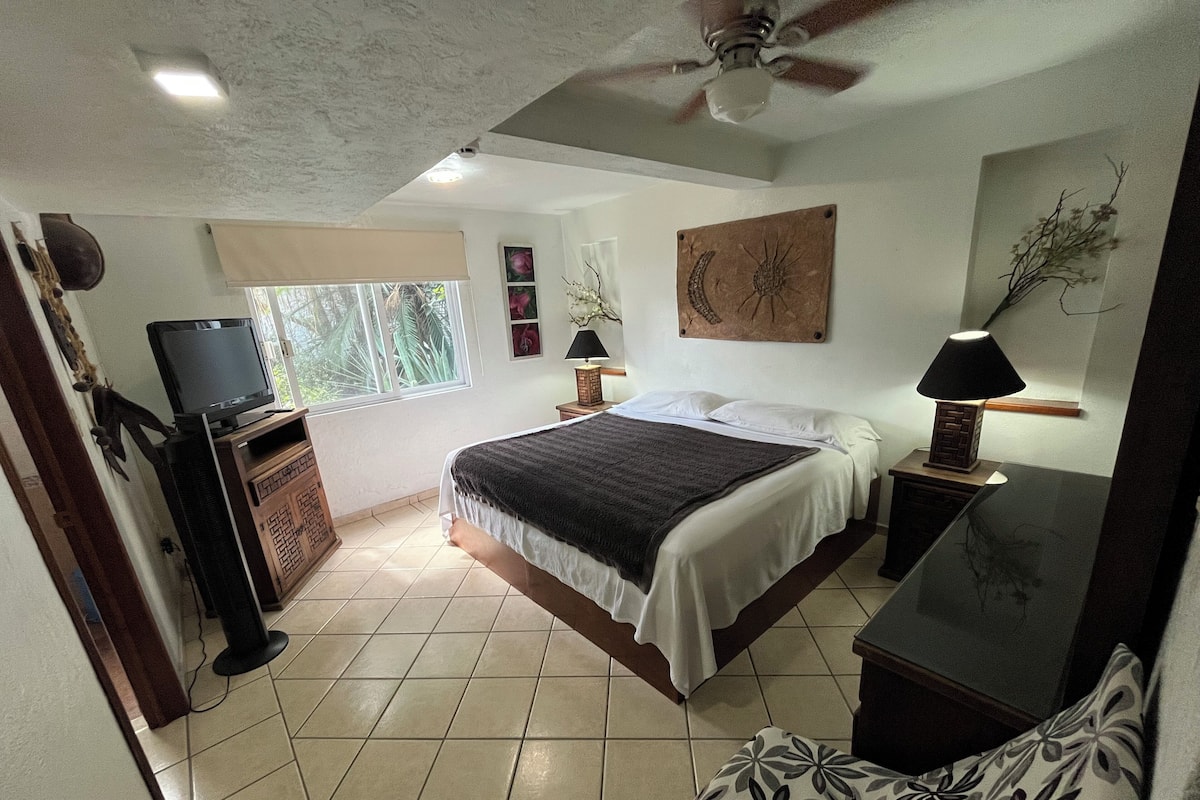 A bedroom with a bed, a television, a ceiling fan, and a window with a view of greenery.