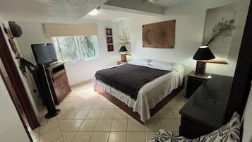 A bedroom with a bed, a television, a ceiling fan, and a window with a view of greenery.