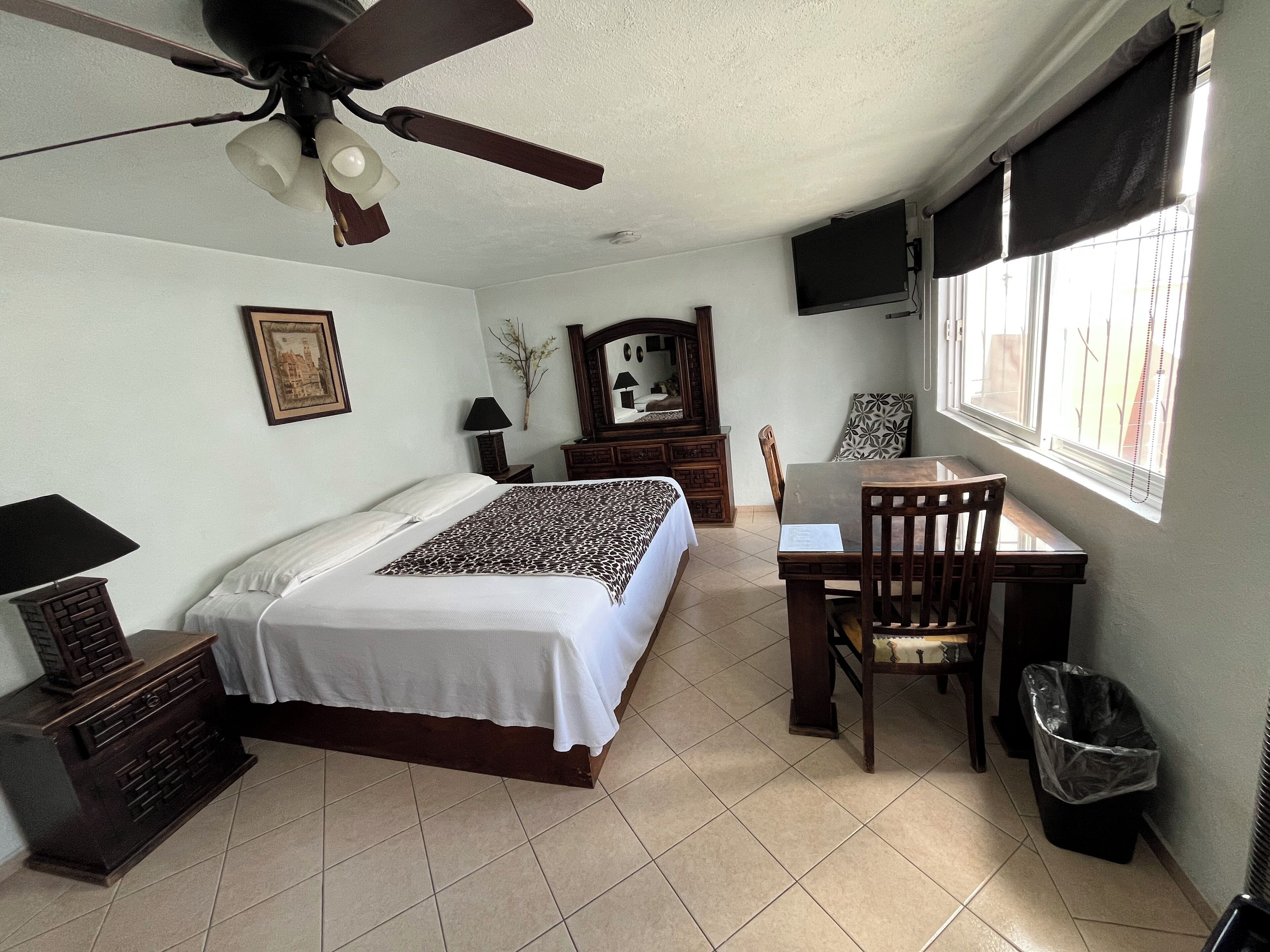 A bedroom with a bed, a desk, a chair, a TV, a ceiling fan, a window, and a trash bin.