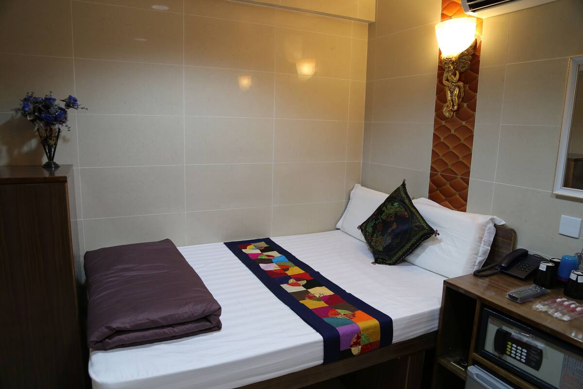 A hotel room with a single bed, a bedside table with a phone and remote, a nightstand, and a wall-mounted light fixture.