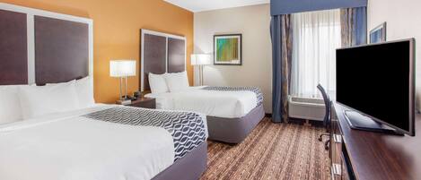 A hotel room with two beds, a desk with a flat-screen TV, and a large window with curtains.