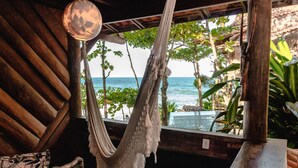 A hammock hanging by a window overlooking the ocean, with a wicker chair and a wooden table nearby. - Pousada Sage Point (Itacaré)