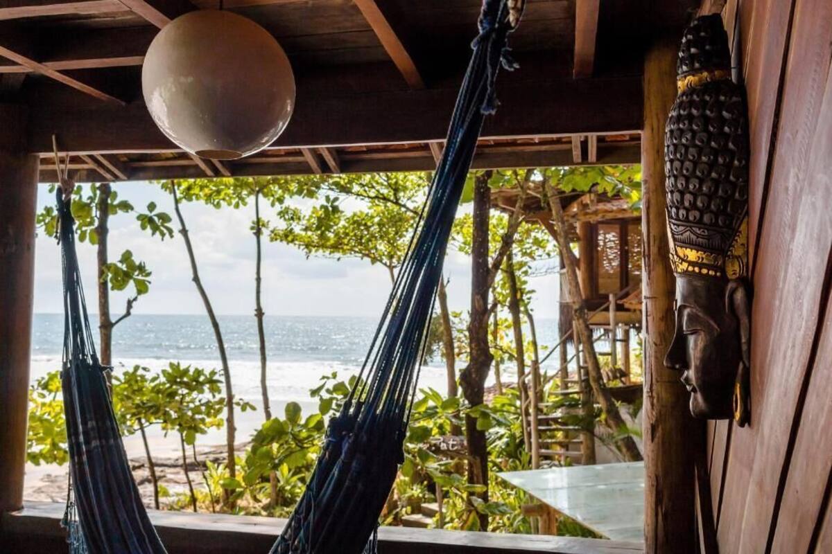 A hammock under a wooden ceiling with a view of the ocean and a tropical garden.
