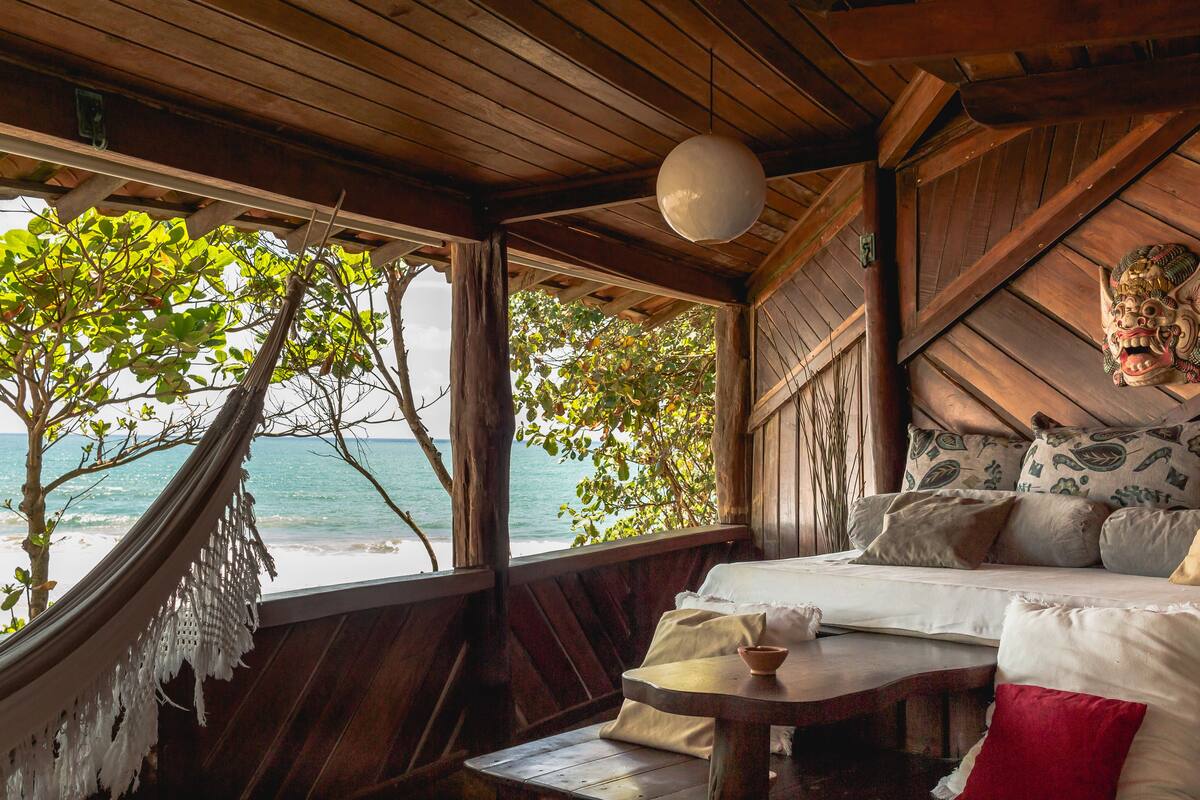 A wooden room with a bed, a hammock, and a view of the ocean.