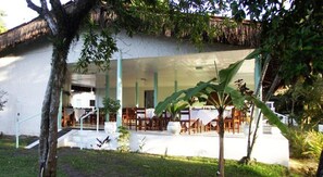 A modern building with a covered outdoor seating area, surrounded by greenery and a thatched roof structure.