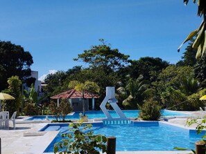 A pool area with multiple swimming pools, a small building with a red roof, and lush greenery surrounding the area.