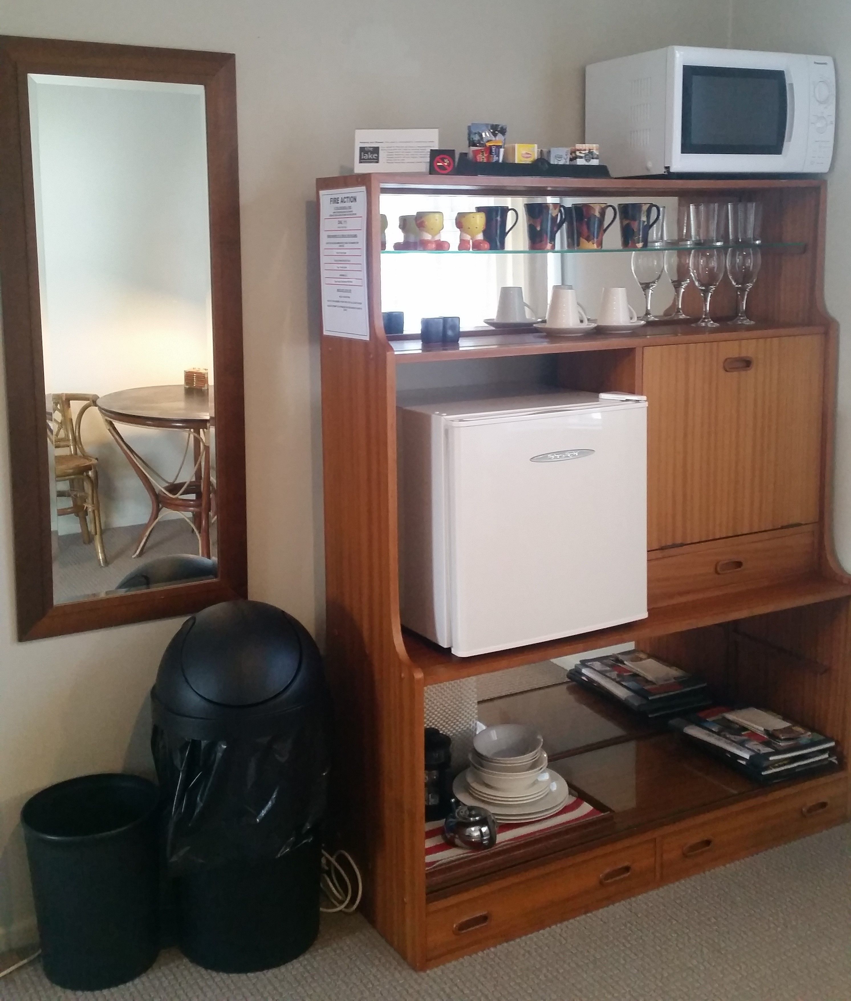 Una habitación compacta con un mueble de madera que alberga un microondas, un refrigerador pequeño y repisas con tazas, vasos y copas de vino. Hay un basurero y una bolsa negra en el piso.