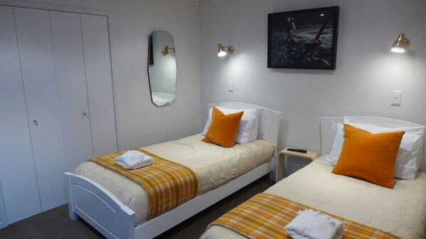 A hotel room with two single beds, each with an orange pillow and a white blanket. There is a white wardrobe, a bedside table with a lamp, and a painting on the wall. - The Lake Motel (Taupo)