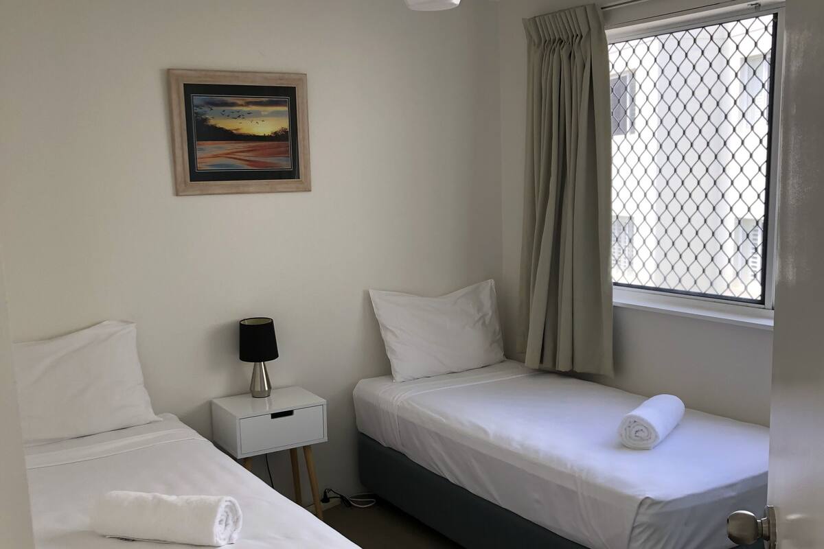 A hotel room with two single beds, a nightstand with a lamp, a ceiling fan, a painting on the wall, and a window with curtains.
