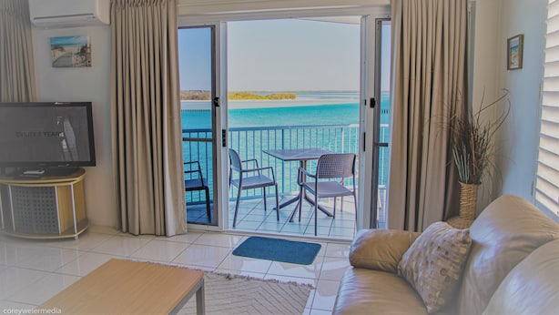 A living room with a television, a sofa, a coffee table, and a balcony with a view of the sea. - Tripcony Quays Apartments (Caloundra)