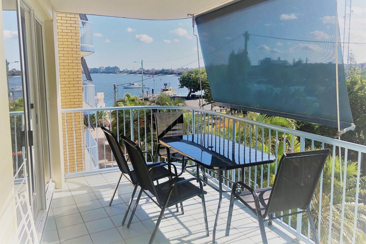 A balcony with a table and chairs, overlooking a body of water with boats and a distant cityscape.