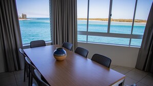 A dining area with a wooden table and six chairs, a decorative bowl, and a view of the ocean through large windows. - Tripcony Quays Apartments (Caloundra)