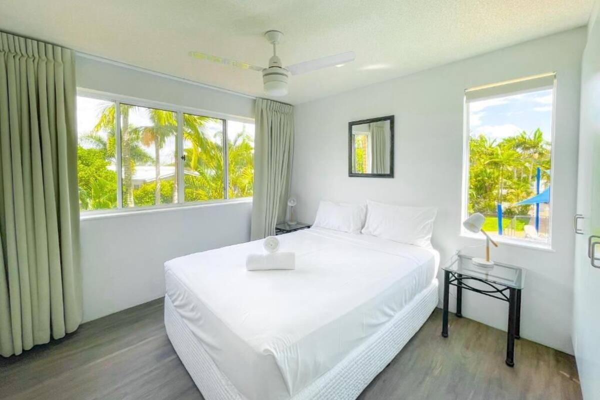 A modern hotel room with a large bed, a ceiling fan, and a view of palm trees through the windows.