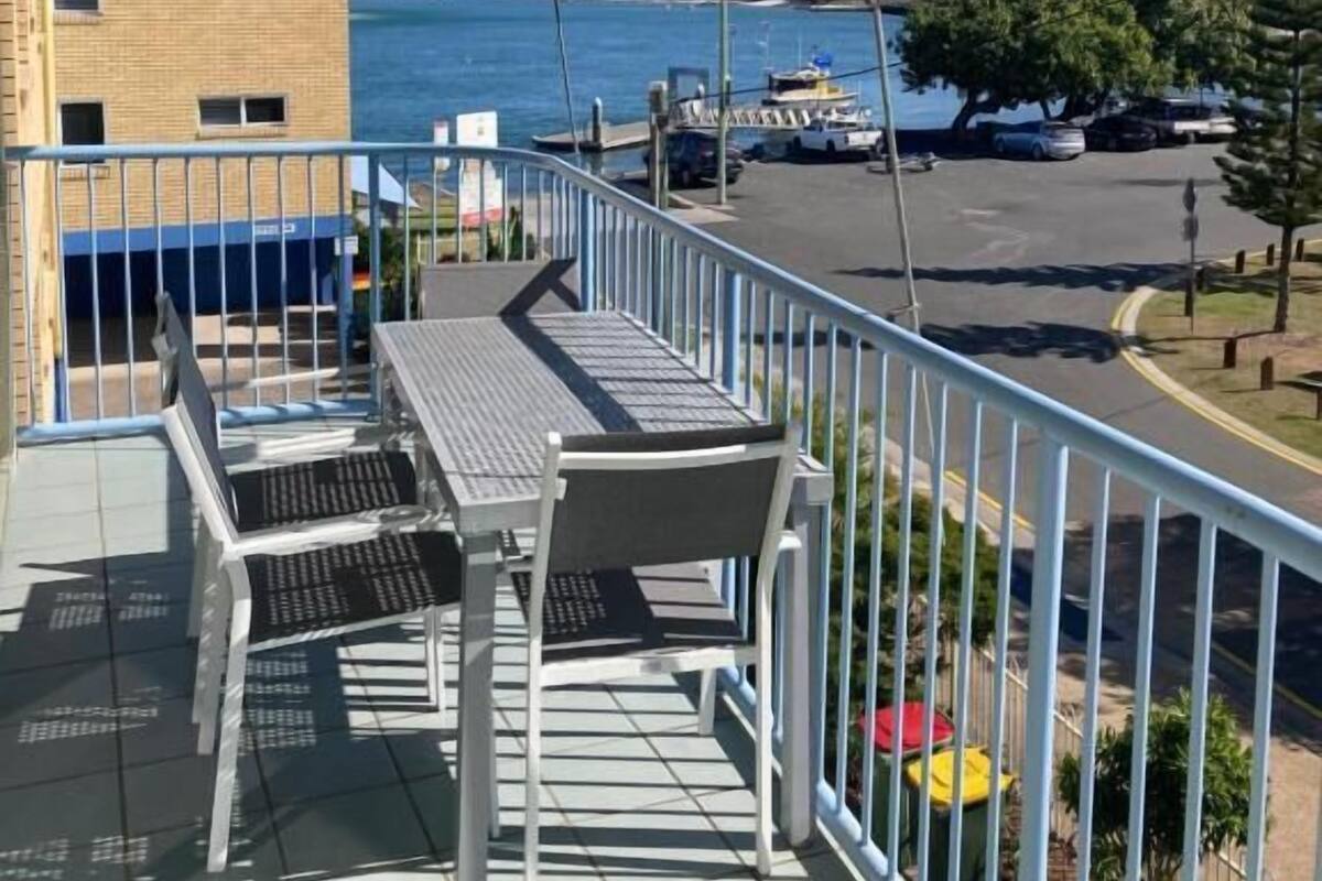 A balcony with a view of the sea, a building in the background, and a parking area.