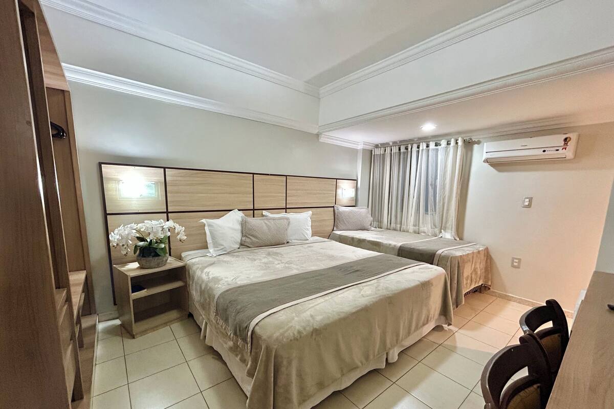Habitación de hotel con dos camas, una mesita de noche con un jarrón de flores, una silla y un equipo de aire acondicionado en la pared.