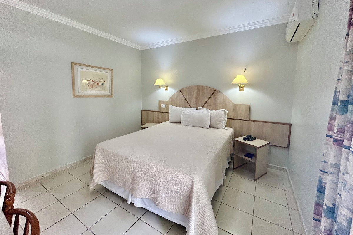Una habitación de hotel con una cama grande, dos mesitas de noche, una lámpara de pared, un cuadro enmarcado y una luz de techo.