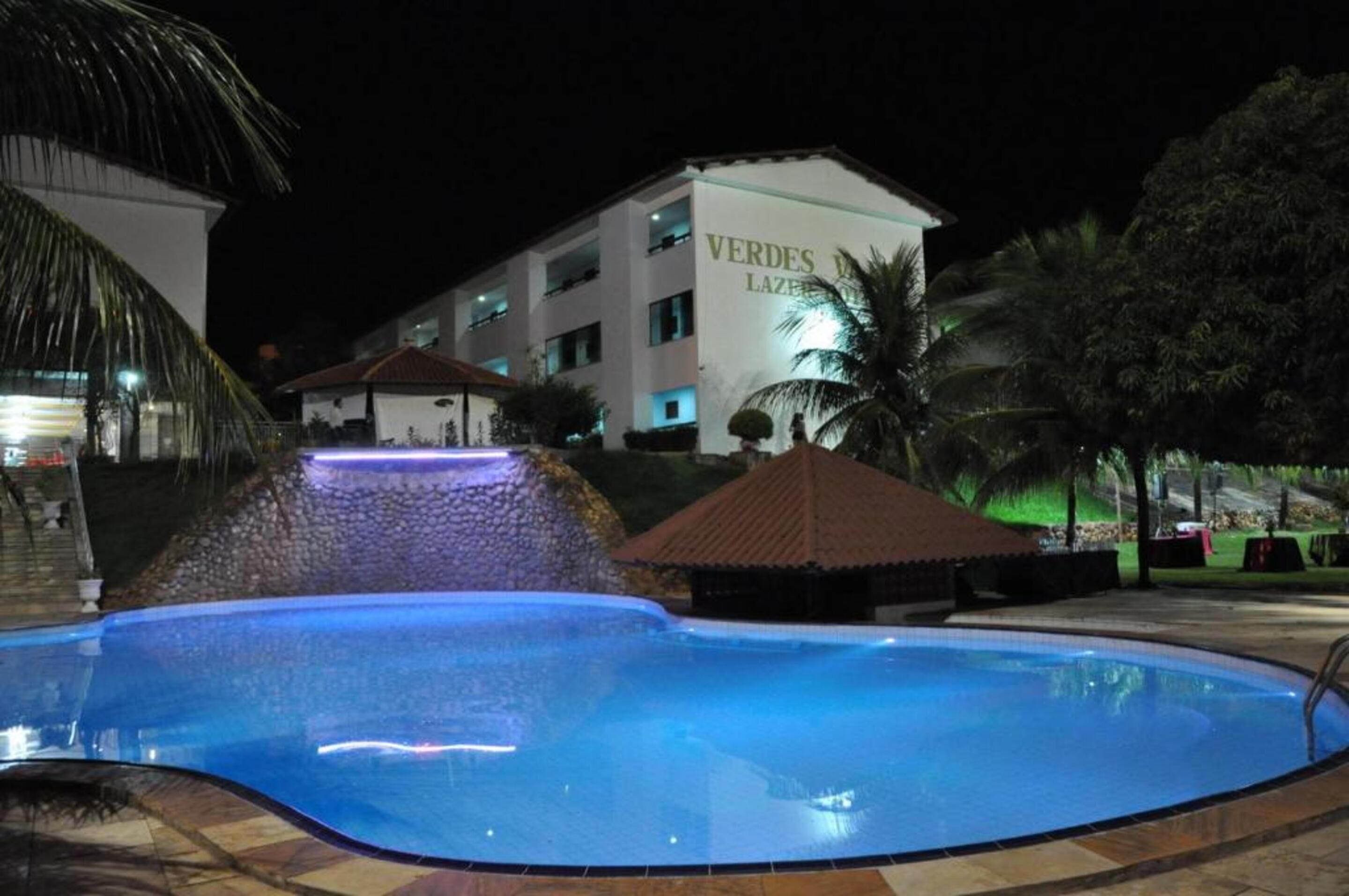 Una piscina con caída de agua, rodeada de palmeras y un edificio con el letrero "VERDES LAZARETA".