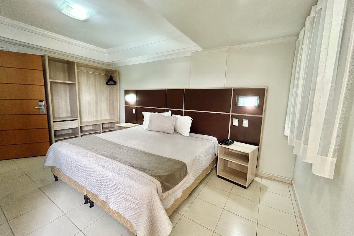Habitación de hotel con una cama grande, un armario, una mesita de noche con teléfono y una ventana con cortinas.