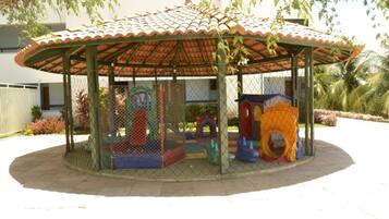 A fenced playground with a tiled roof, surrounded by a paved area and palm trees.