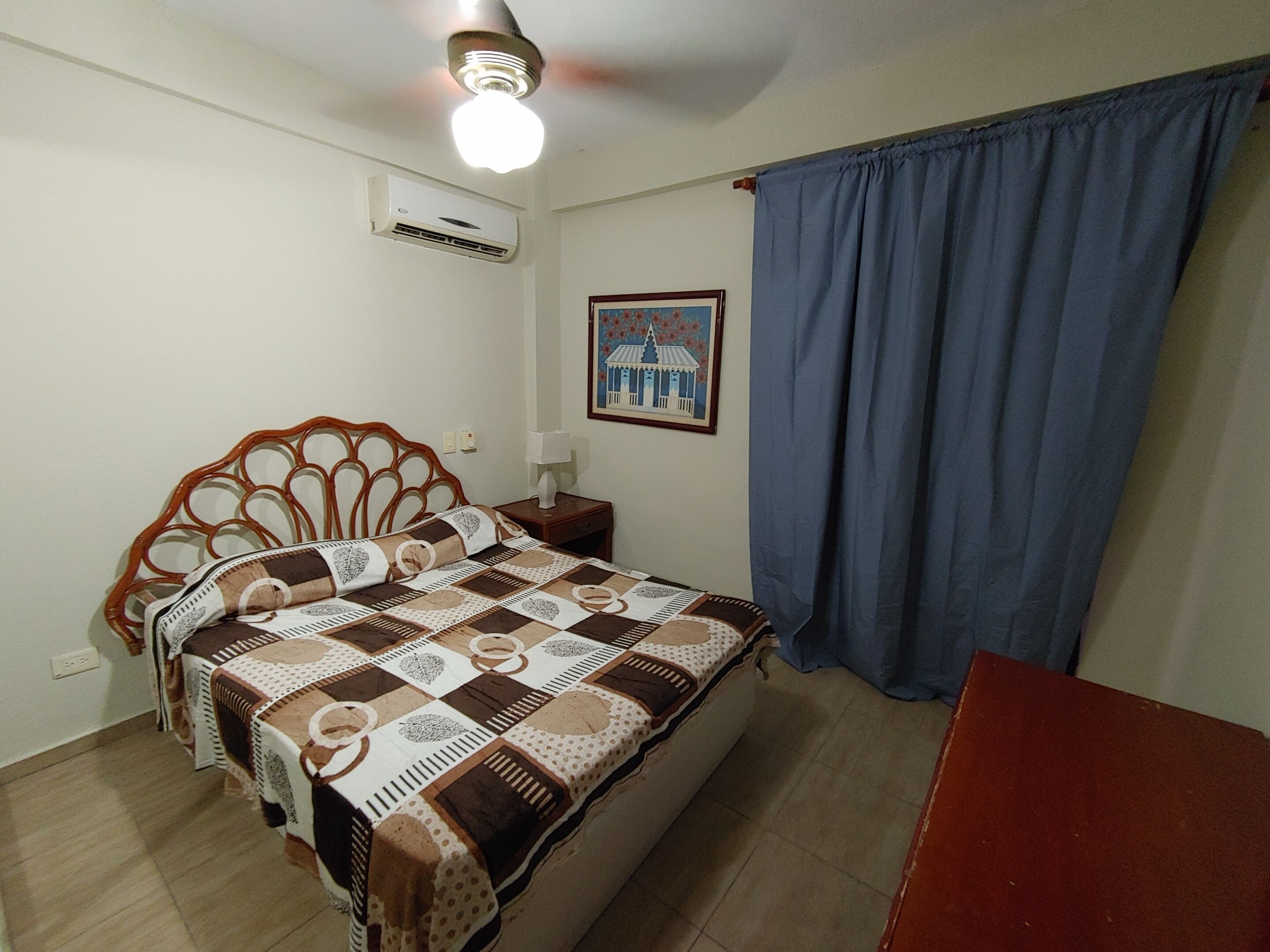 A bedroom with a bed, a nightstand, an air conditioner, a painting, and a window with curtains.