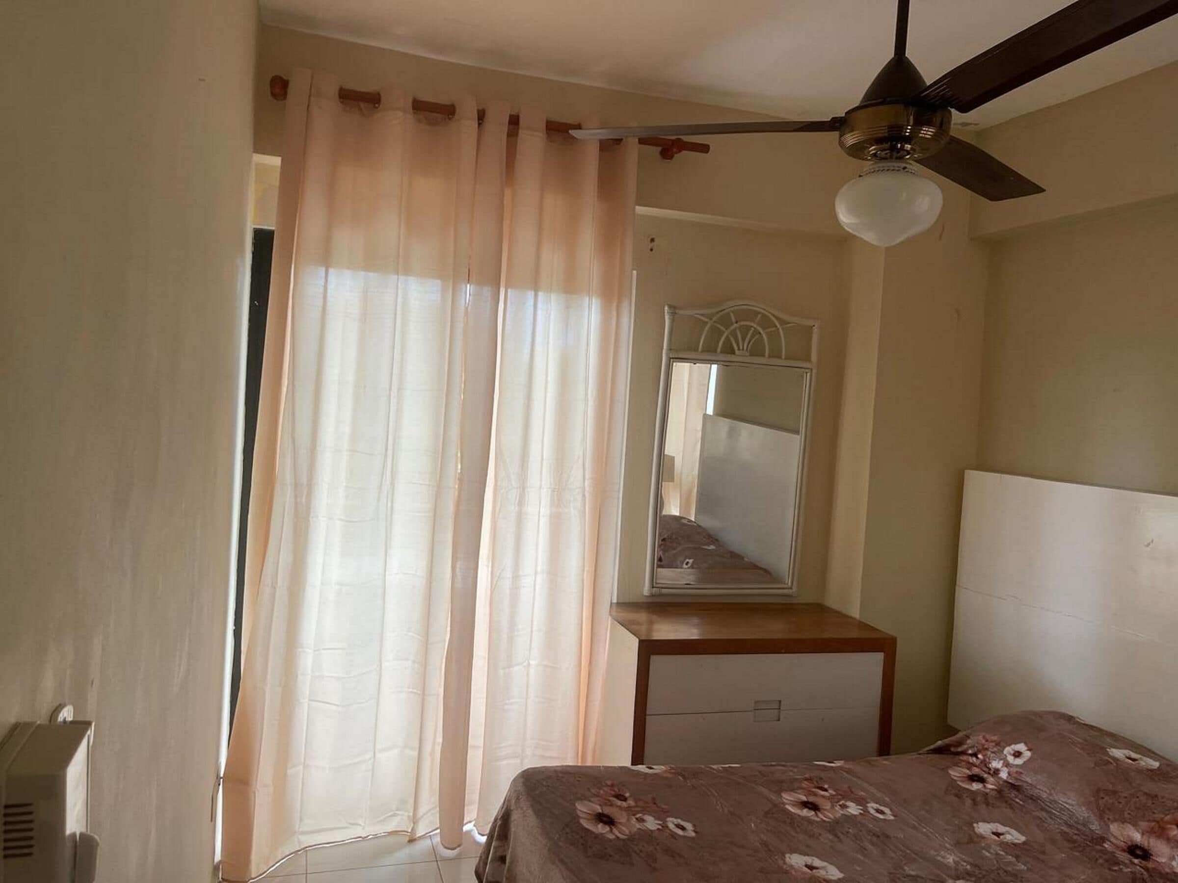 Quarto com cama, mesa de cabeceira, espelho, ventilador de teto e janela com cortinas.